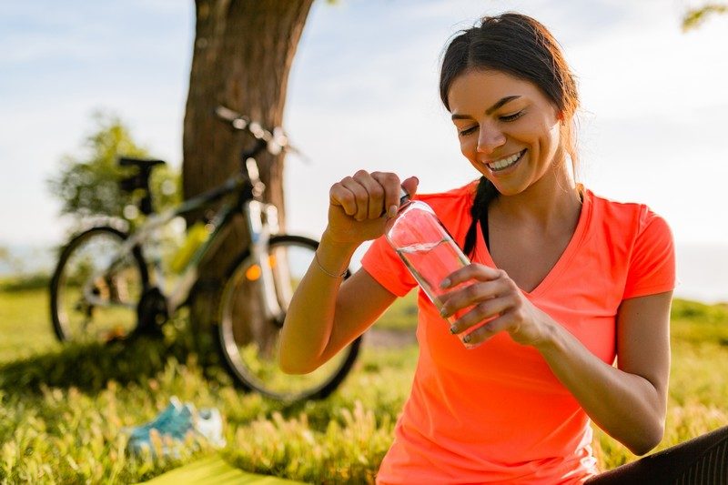 smiling beautiful woman drinking water in bottle doing sports in morning in park nature doing yoga on mat, pink fitness outfit, happy healthy lifestyle, music in earphones, bicycle on background